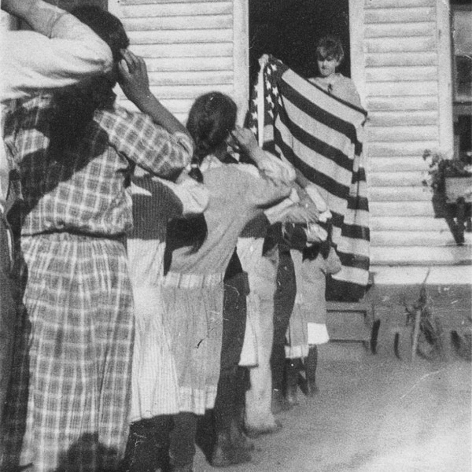 A black and white photo of a teen-aged boy holding an American flag while standing in a doorway of a white building as a line of children salute the flag. children, a flag, and school house.