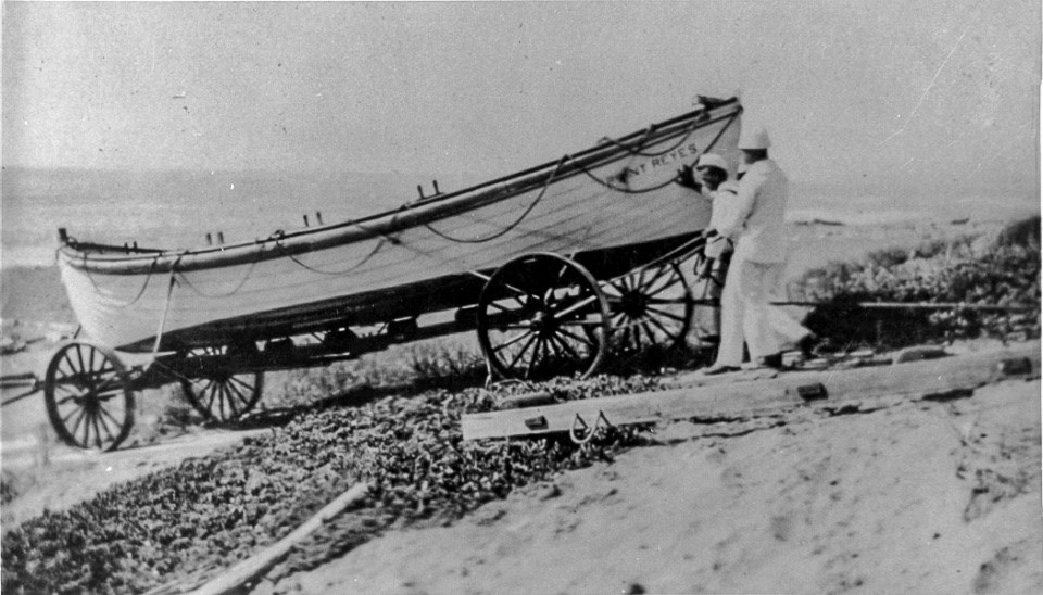 A black and white photo of two men in white uniforms maneuvering an approximately 25-foot-long open-top surfboat on a wheeled cart.