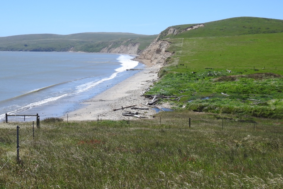 A black and white photo of a garage-like structure and a surf boat adjacent to the beach to the left with cliffs in the background.