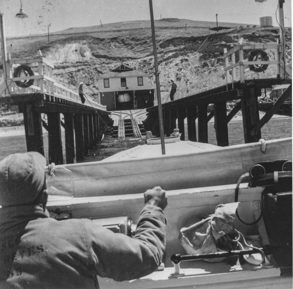 A black and white photo of a man on the left at the helm of a boat as it is guided up a ramp to a boathouse.