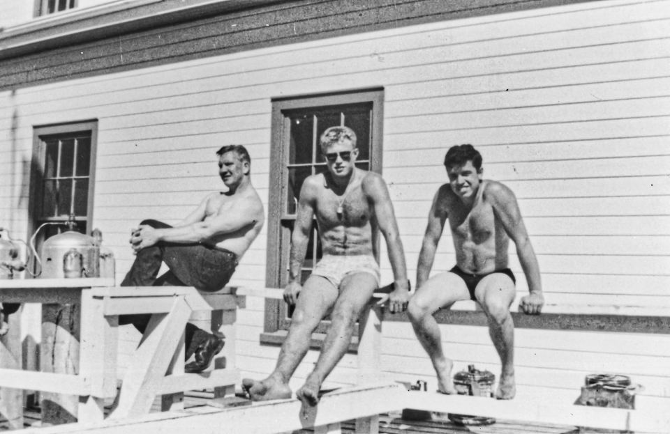 A black and white photo of three men sitting on railing of a white building..