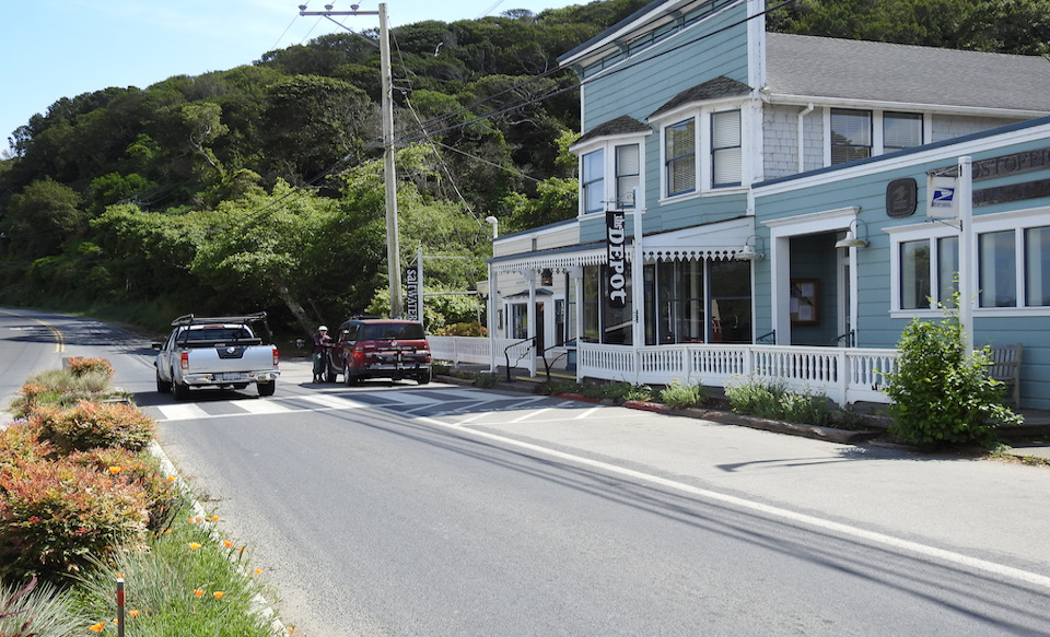 A black and white photo of eleven young men running a race on a dirt road past with a crowd of people and buildings in the early 1900s.