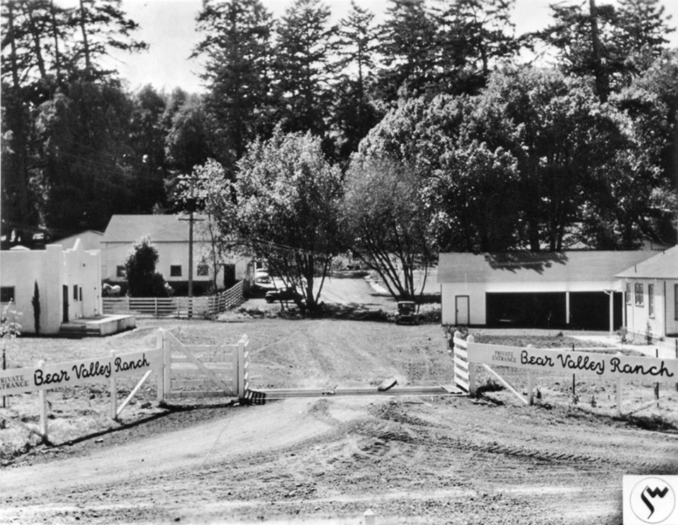 A black and white photo of dirt driveway passing over a cattle guard. Painted on white fences on both sides of the driveway is "Private Entrance. Bear Valley Ranch." Beyond the cattle guard are a number of white buildings, beyond which are tall trees.