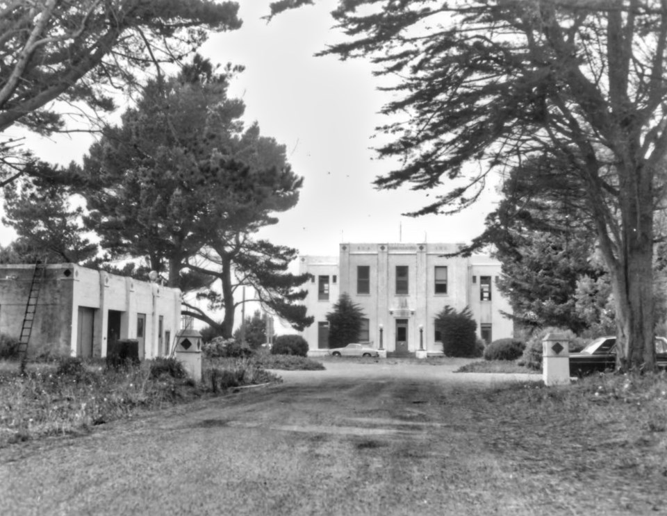 A black and white photo of two white, Art Deco-style buildings, trees lining a driveway, and two 1970s-era cars.