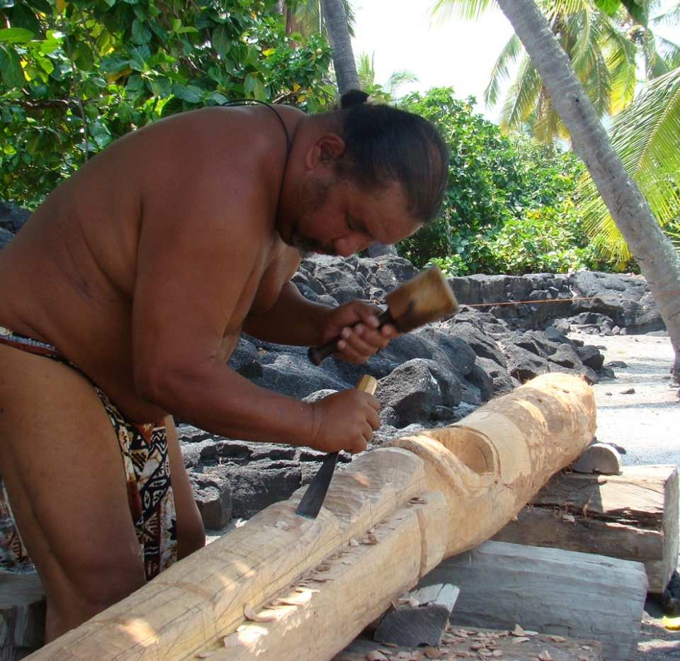 Historic photograph of a man carving a kiʻi in front of a rock wall