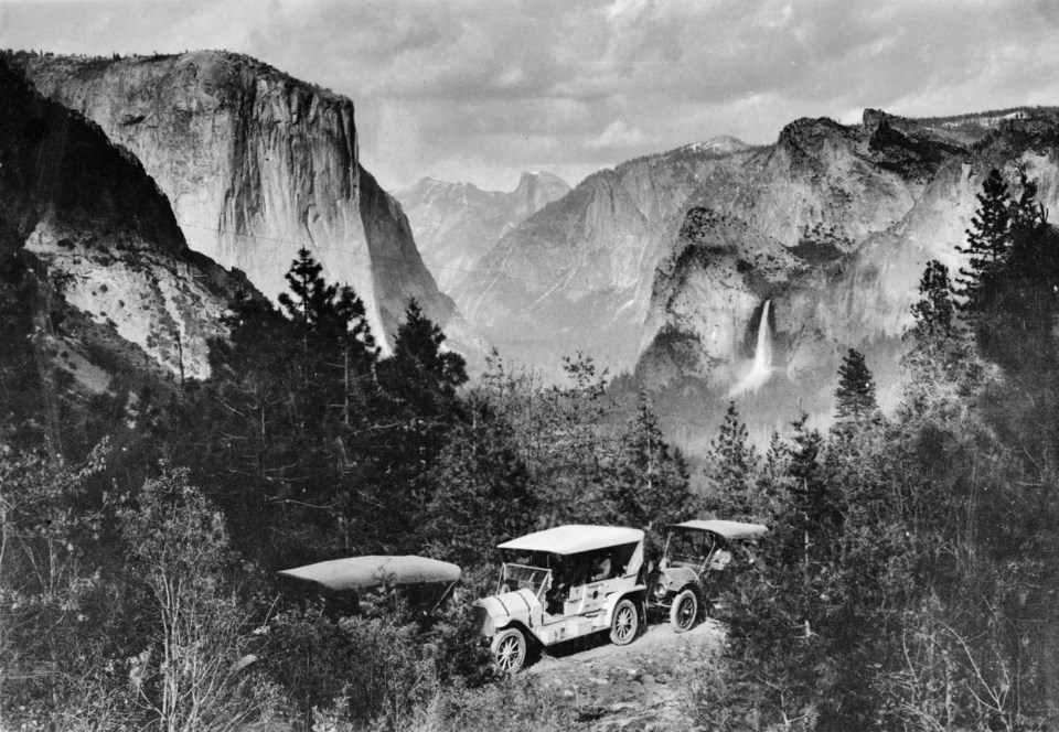 Several older cars in a row on a road with cliffs and a waterfall in background.