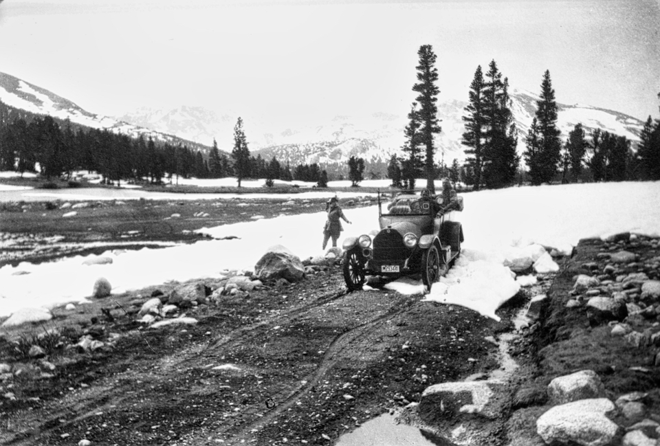 An older car on a road with some snow, trees, and a frozen meadow.