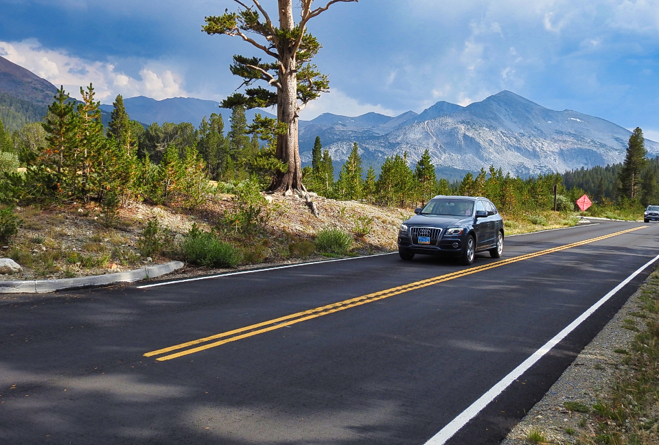 An older car on a road with some snow, trees, and a frozen meadow.