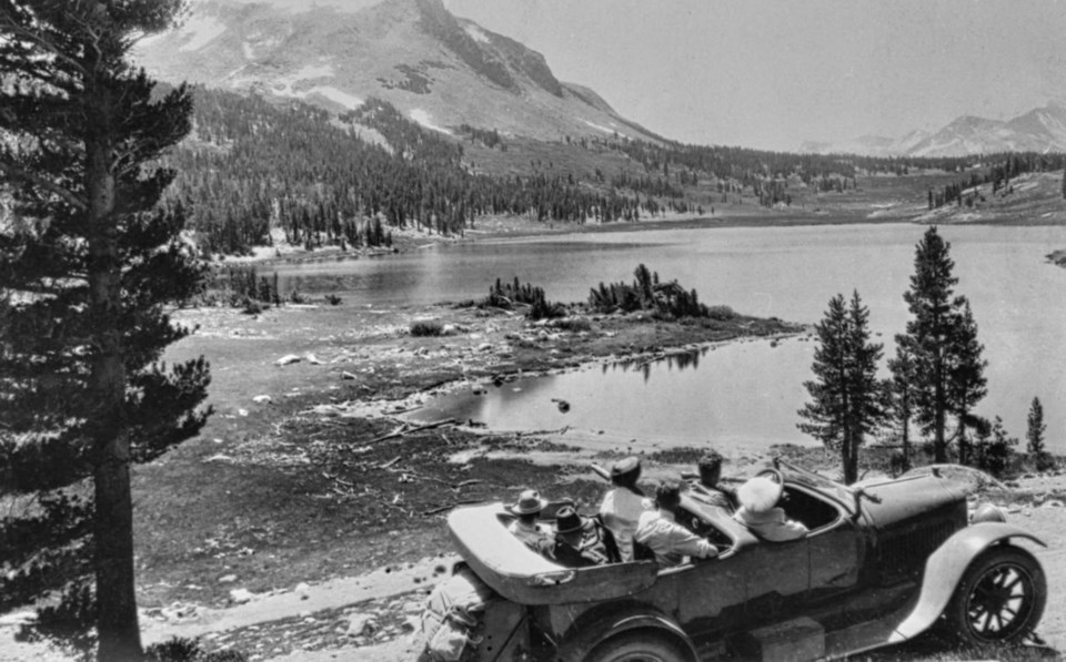 An older convertible car with several people look at a lake with mountains in background.