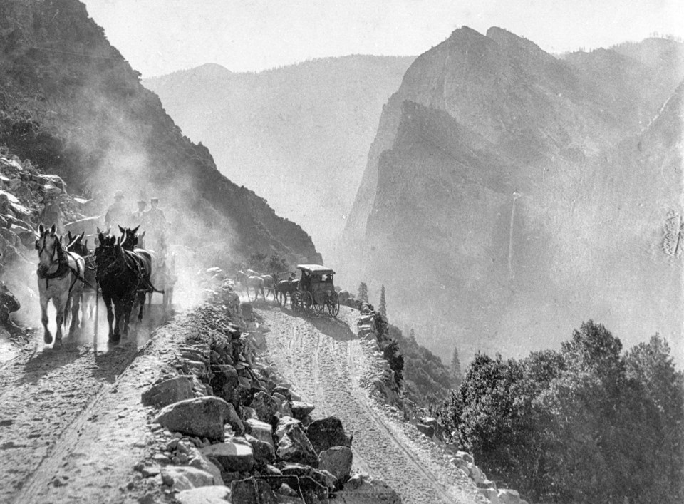 Horse-drawn stages on a road cut into the edge of a hill with cliffs in the background.