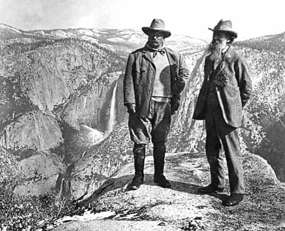 Two men stand on a rock with waterfalls and mountains in background.