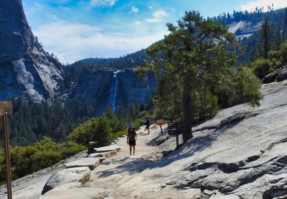 Two women sit on a granite slab next to a trail with waterfalls in the background.