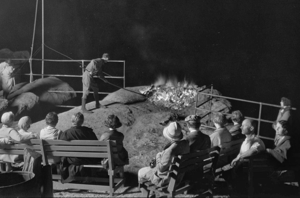A group of people watch a man tending a fire next to a cliff at night.