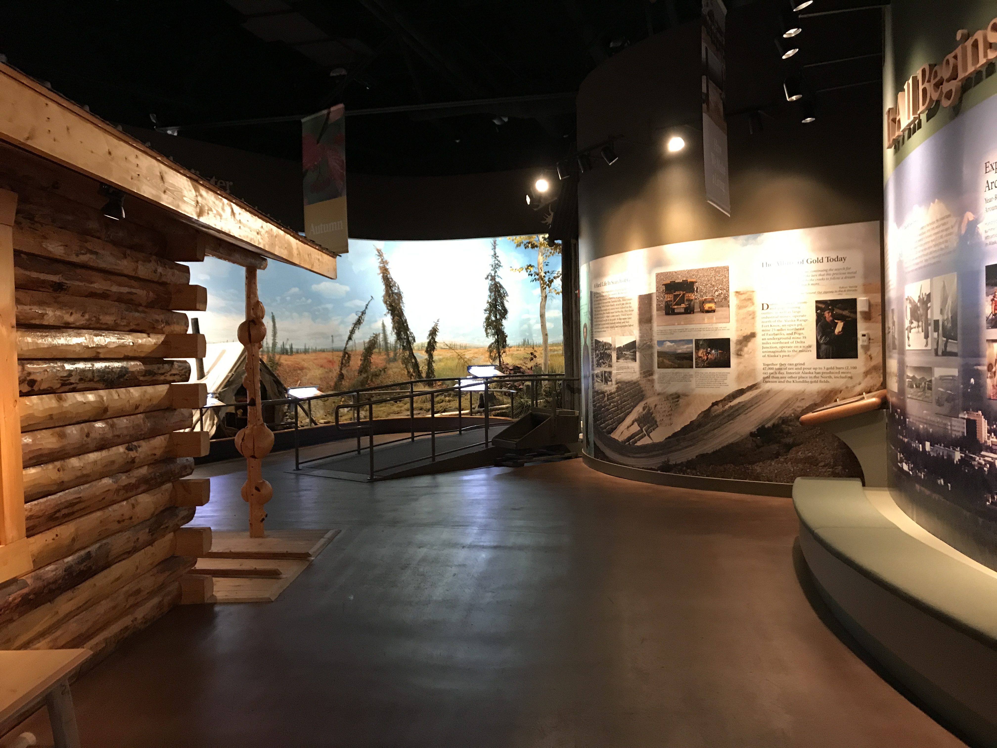 Outdoors; flower beds in forefront to left of cement sidewalk leadng to a large wooden block building.