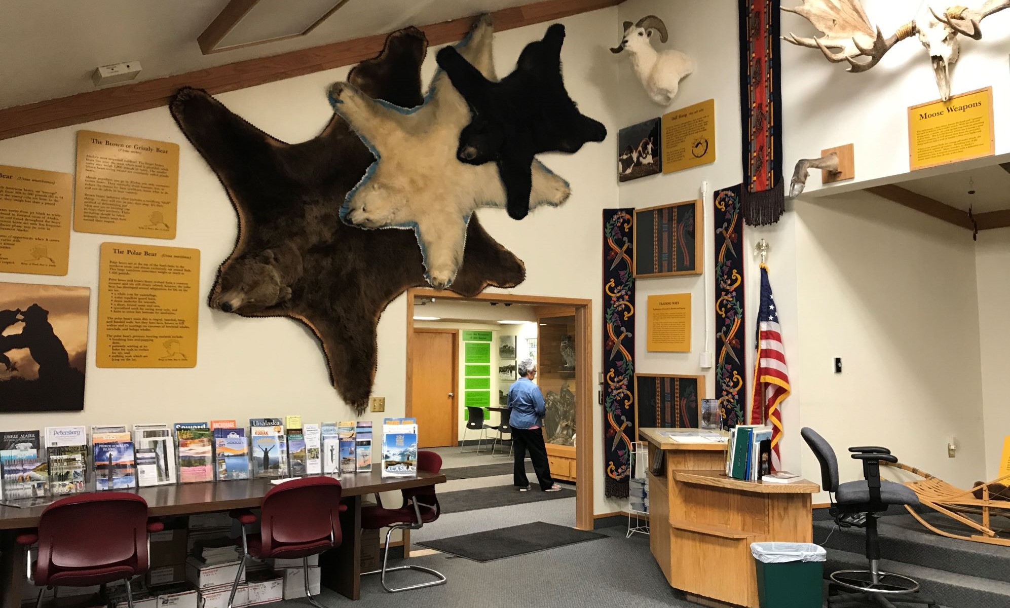 Outdoors; wooden building on left, 2 flags and 4 wooden signs in middle foreground, sky above and grass below.