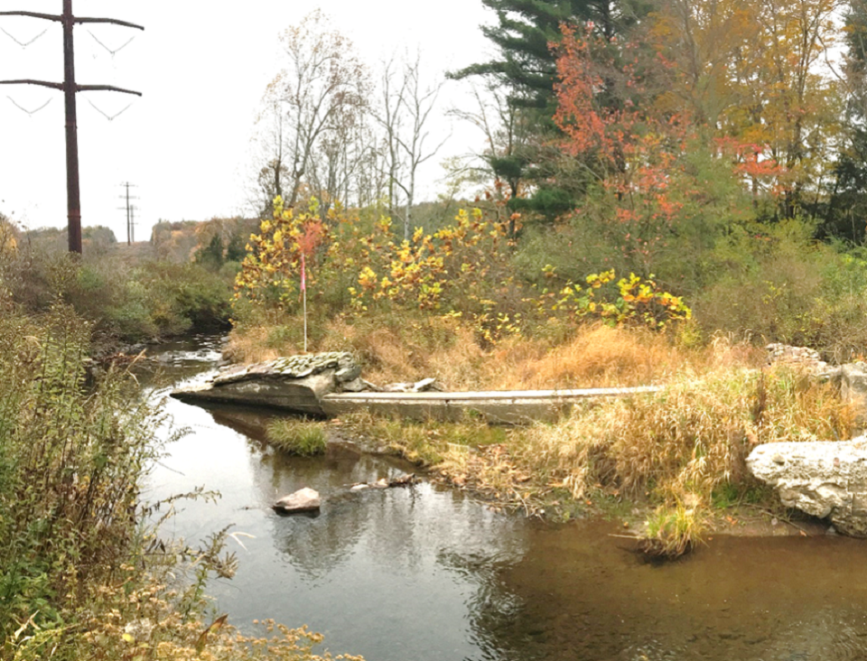 A stream with a crumbling concrete dam in the foreground. The background shows power lines and trees with green, red, and yellow leaves.