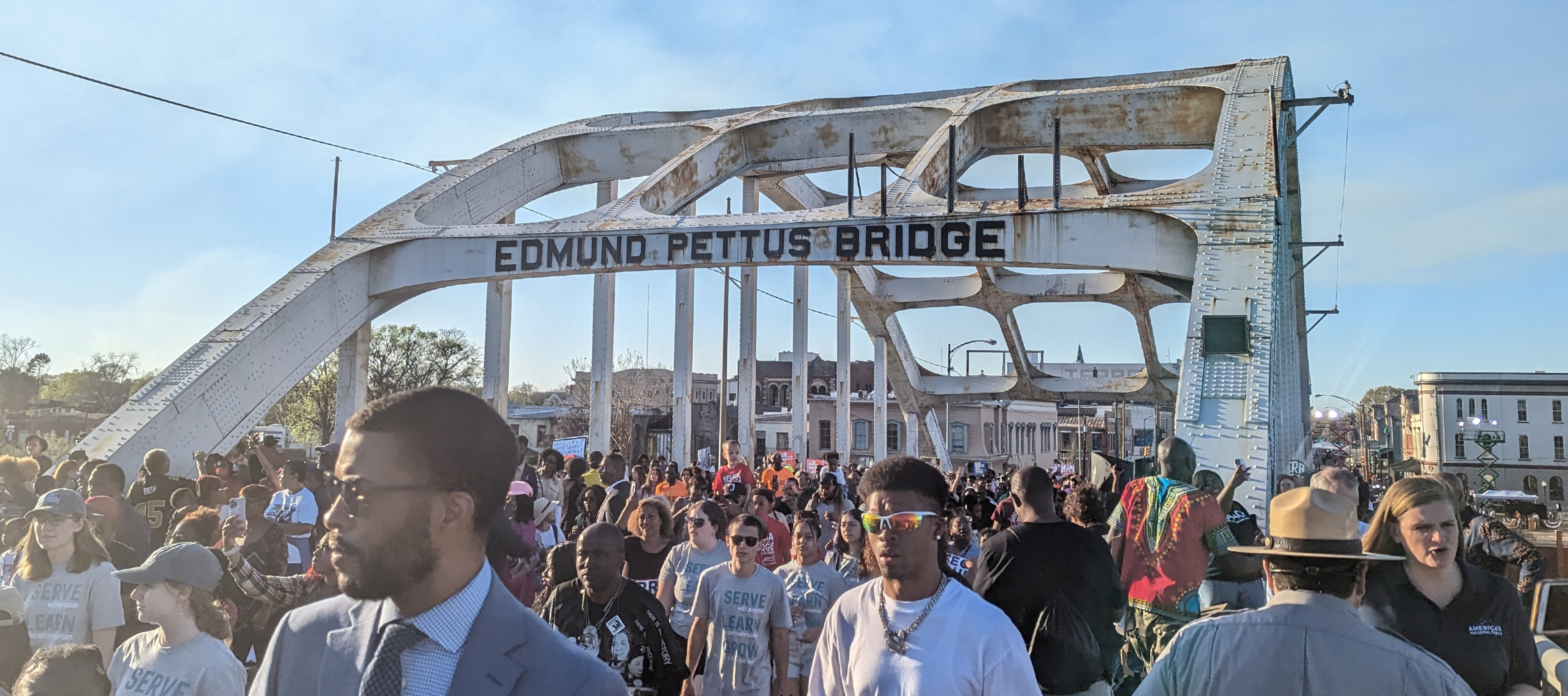 A crowd walking across a bridge, the arched steel beams overhead the people read, "Edmund Pettus Bridge,” blue sky in the background.
