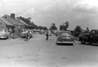 People and cars gather along a paved parking area in front of a low building.
