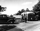 Two people, facing each other, stand at the roadside between a trailer and a 1940s-style car.
