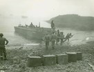 Uniformed soldiers carry weapons and supplies ashore on a rocky beach from a boat.