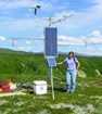 A woman with long, dark hair, lilac shirt and jeans stands next to a climate monitoring station.