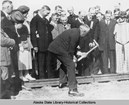 man hammering a ceremonial spike on a railroad, surrounded by people