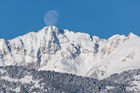 Electric Peak is covered with snow below a full moon on a blue sky day.