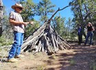 A man stands talking to a group of people near a structure made out of tree limbs.