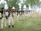 Men in 18th Century-style soldiers uniforms stand shoulder to shoulder firing muskets. 