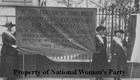 Three women with suffrage sashes stand outside the White House holding a large banner 