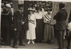 Two suffragists carrying banners held by the arms by a police woman as a crowd looks on