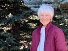 Smiling older woman with white hair, white shirt and pink jacket in front of green trees.
