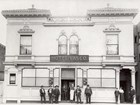 10 people stand in front of 2-story Spanish style hotel. Sign on central door reads 