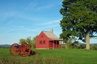 A red, two-story clapboard house sits on a verdant grass field near a line of trees and shrubs.
