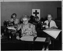 Three senior women use sewing machines to work on white squares of fabric as Red Cross volunteers