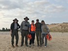 A group of five people stand on a beach. Three are NPS employees wearing the uniform