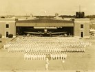 Sepia photo of people in white in formation outside airplane hangar 