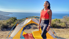 a woman poses next to a tent on a cliffside overlooking the ocean