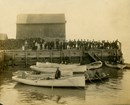 People standing on a wooden dock, below them a man stands in a small wooden boat.