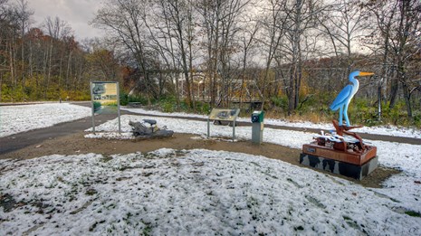 Two graphic panels, statue of eagle on a log, & tall blue heron statue beside paved trail in snow.