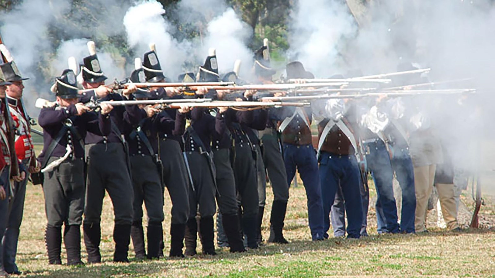 A line of living historians dressed in soldiers\' uniforms firing musket rounds.