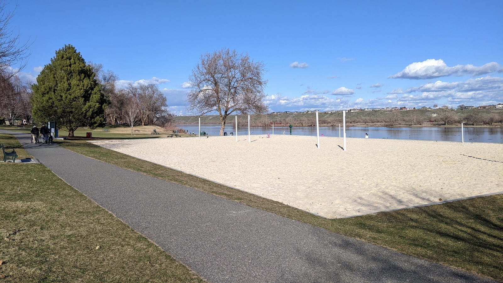 A paved path runs alongside a rectangle of white sand surrounded by a few trees and green grass.  