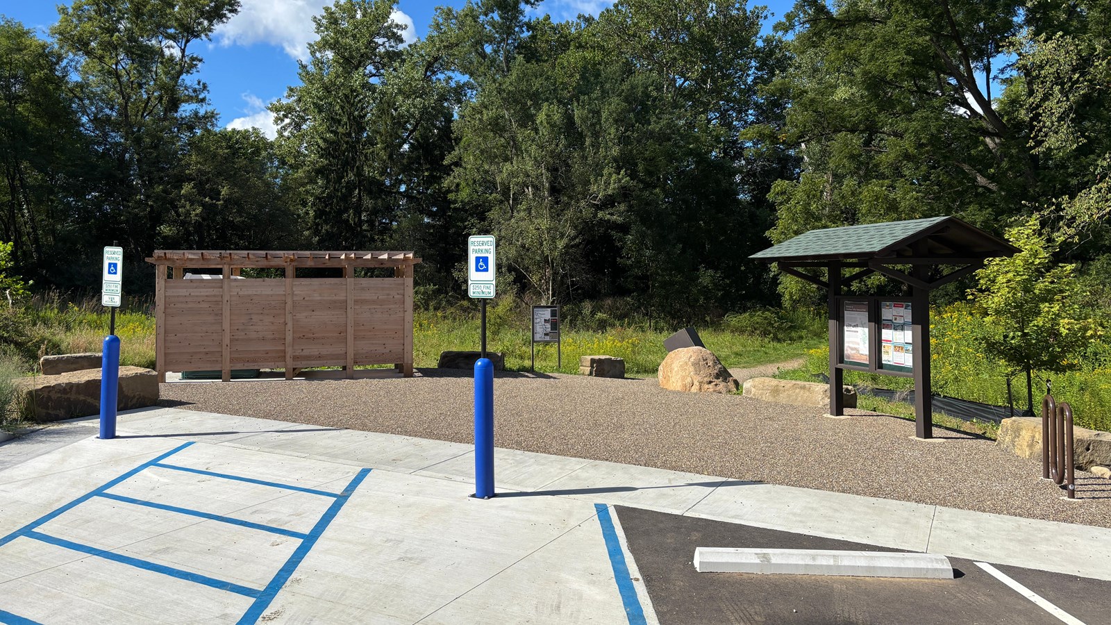 A wooden structure and roofed kiosk near the edge of a parking lot; a trail leads into a forest.