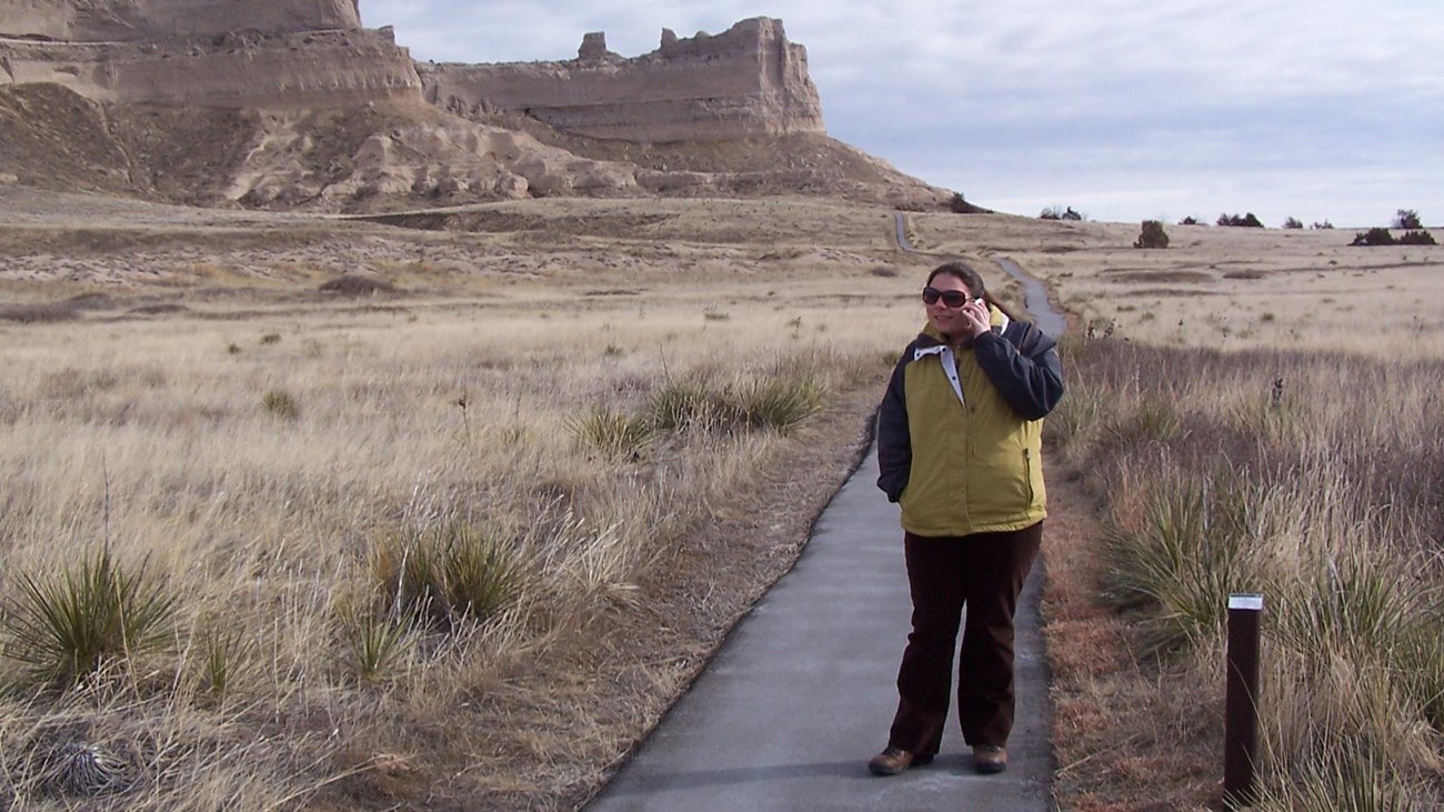 A woman stands on a paved trail with a cellular phone to her ear and a view of sandstone bluffs.