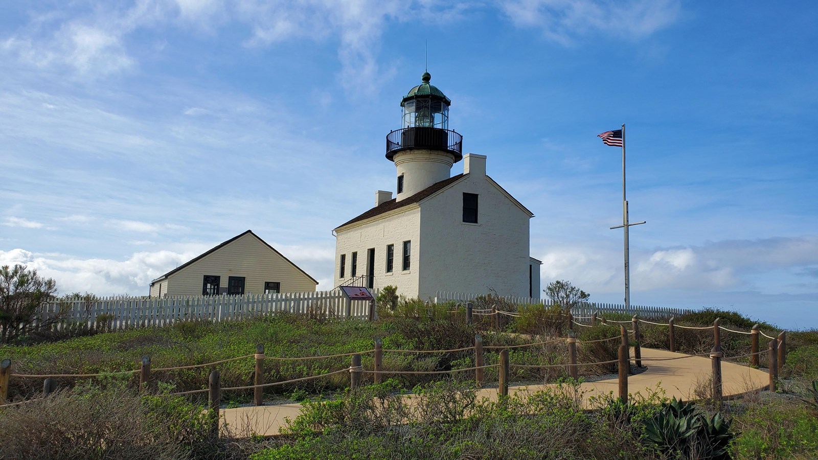 A brown path winds through green plants to a white lighthouse and a small white building.