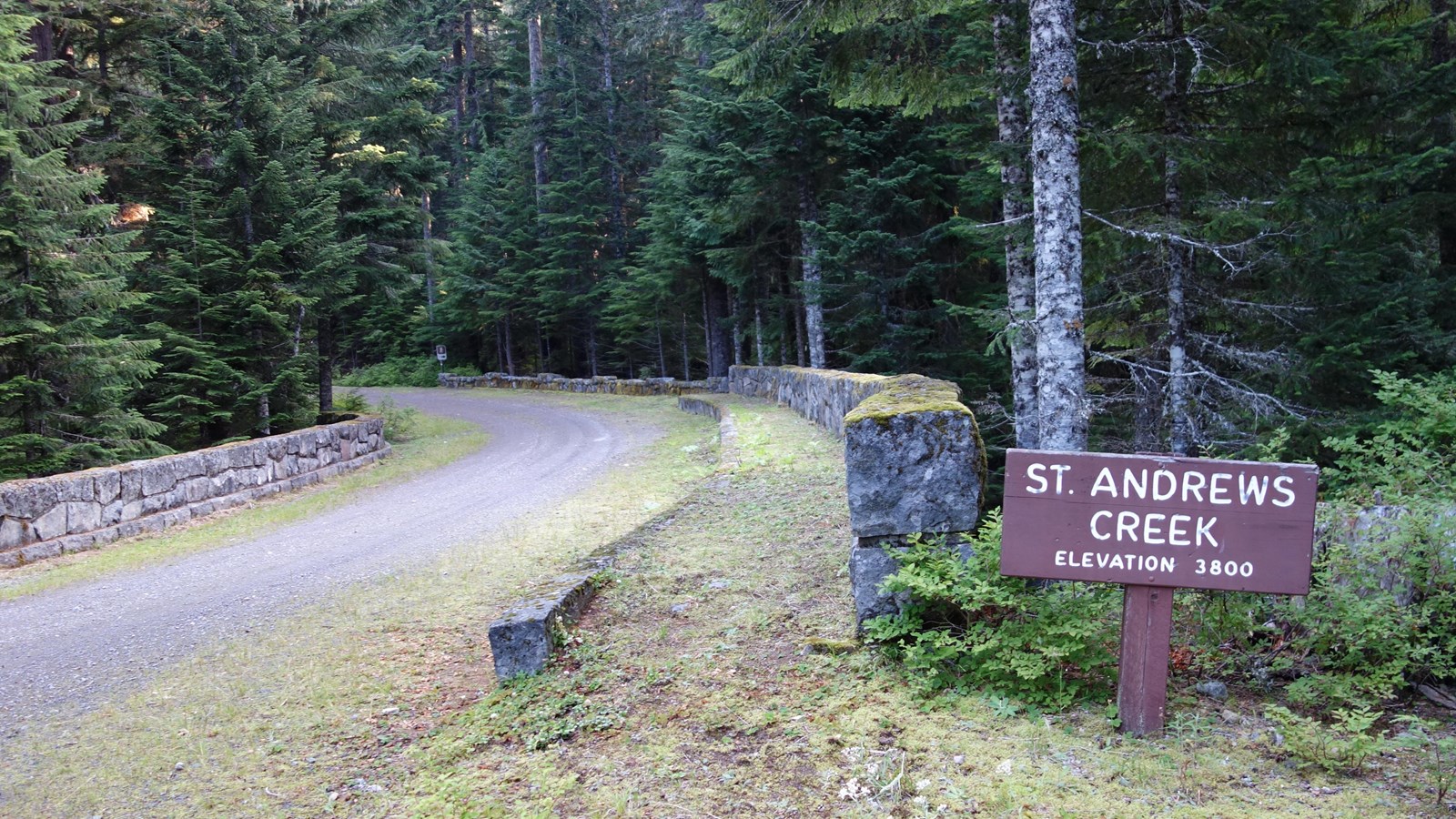 A stone bridge with mossy rock walls. A wood sign next to the bridge reads 