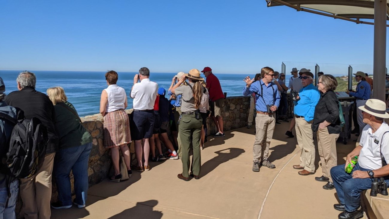 A crowd of people gather under a white, cloth covering. They look over a fence towards blue water.