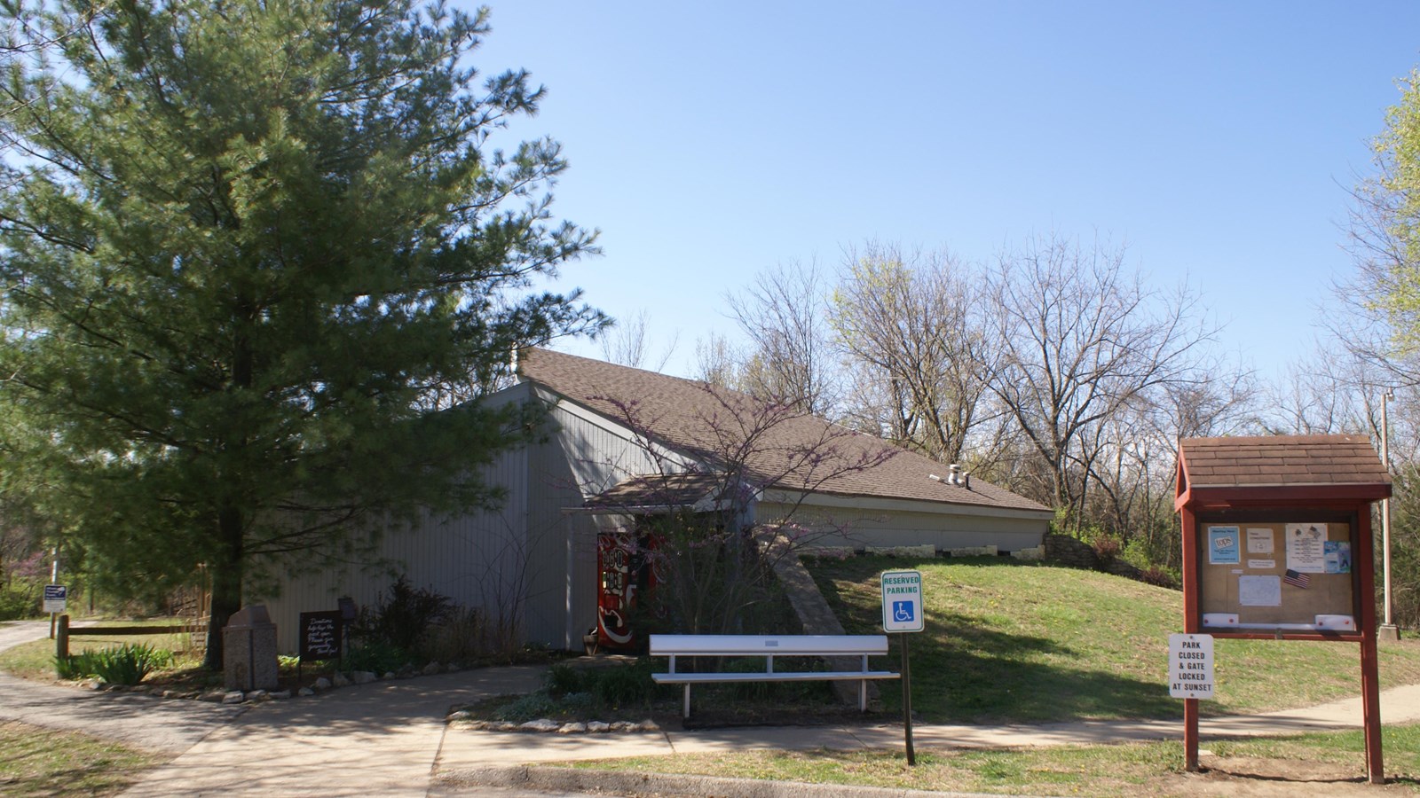 A cement path leads to a slanted-roof building. A large tree and a sign flank the path.