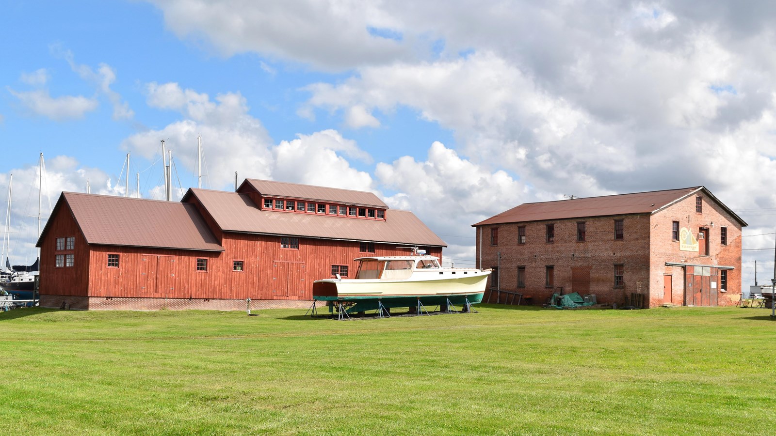 Two large buildings, one wooden and one brick, next to a boat on stands with a grassy foreground.