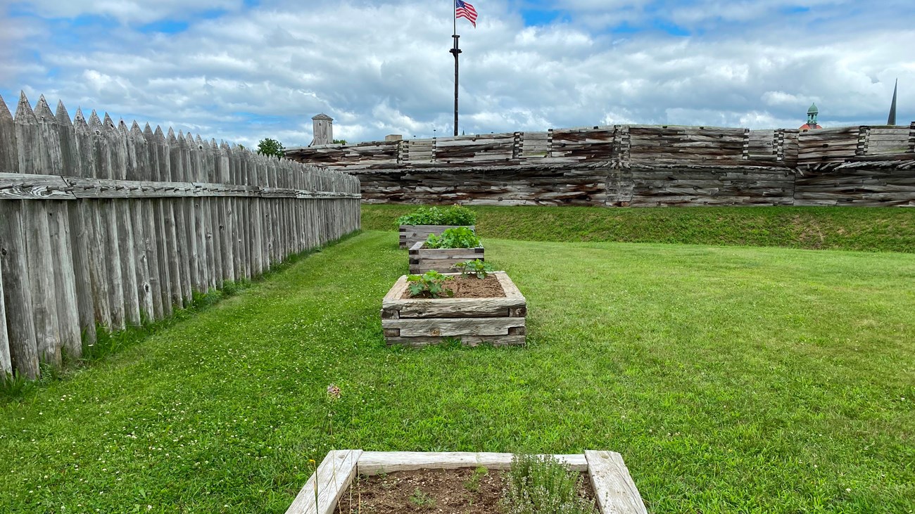 3 raised garden beds in a grassy area in front of the fort wall and inside the picket fence. 